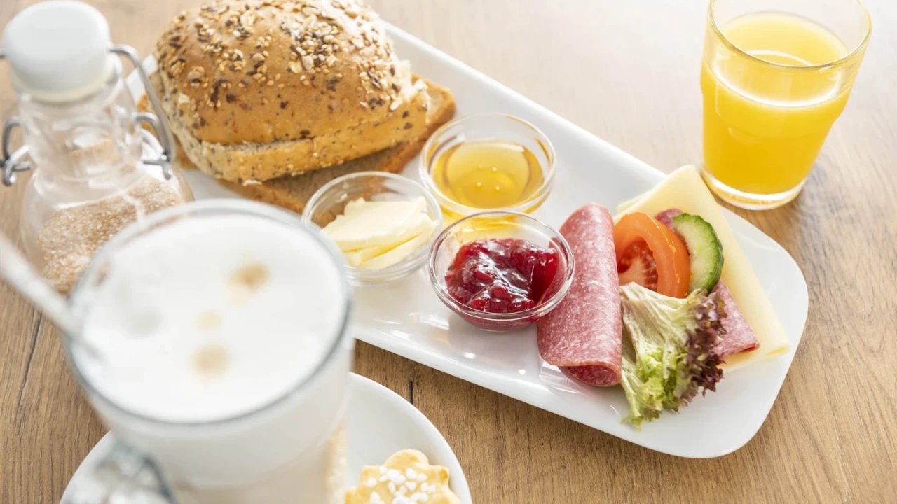 Fr&uuml;hst&uuml;cksteller mit einem Mehrkornbr&ouml;tchen, kleinen Sch&auml;lchen mit Honig, Butter und roter Marmelade, einer Scheibe Salami, K&auml;se und Gem&uuml;segarnitur aus Salat, Gurke und Tomate auf einem wei&szlig;en Tablett. Daneben ein Glas Orangensaft und davor ein Glas Latte Macchiato mit einem Keks.