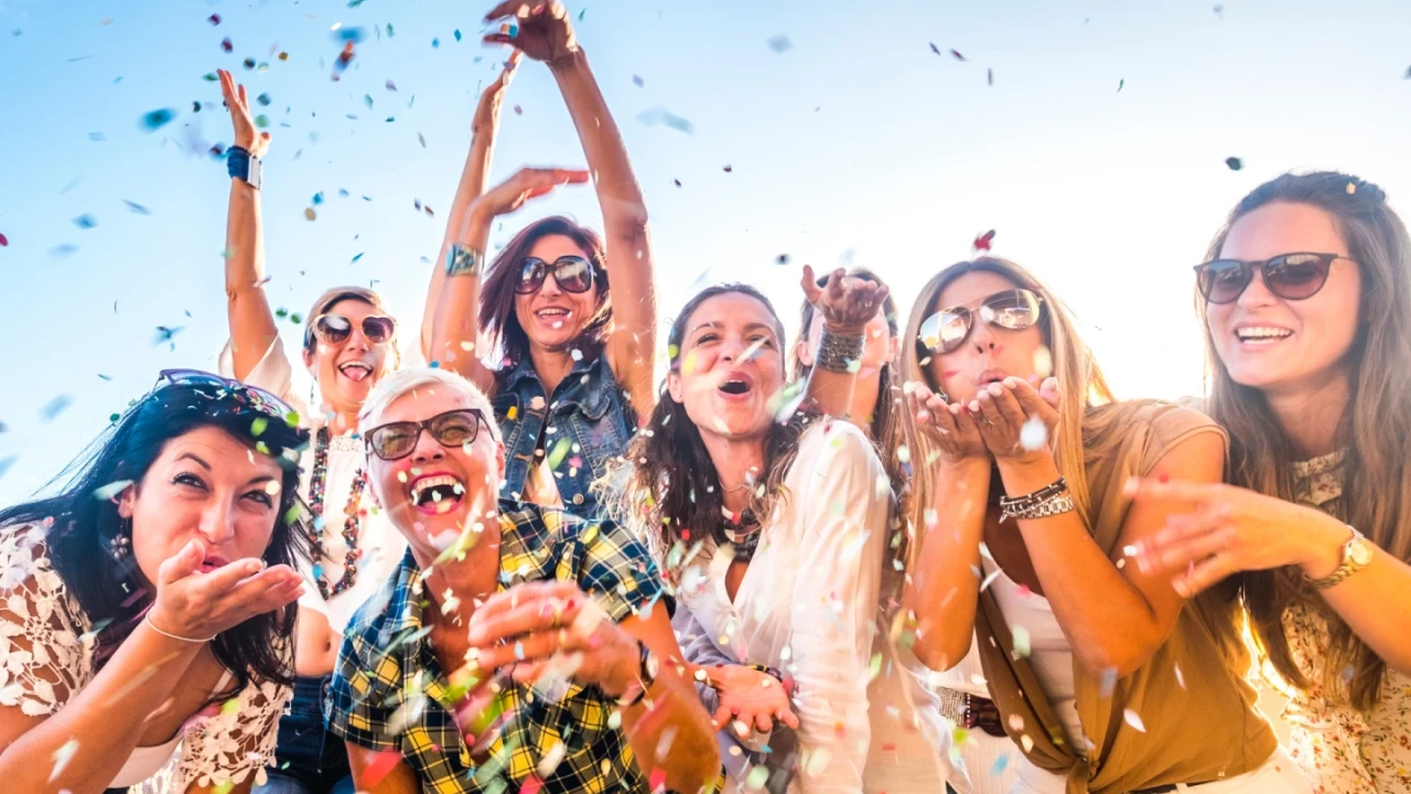 Gruppe von acht Personen, &uuml;berwiegend Frauen, die lachen, feiern und buntes Konfetti in die Luft werfen. Alle tragen Sonnenbrillen oder helle Kleidung, vor einem klaren blauen Himmel.
