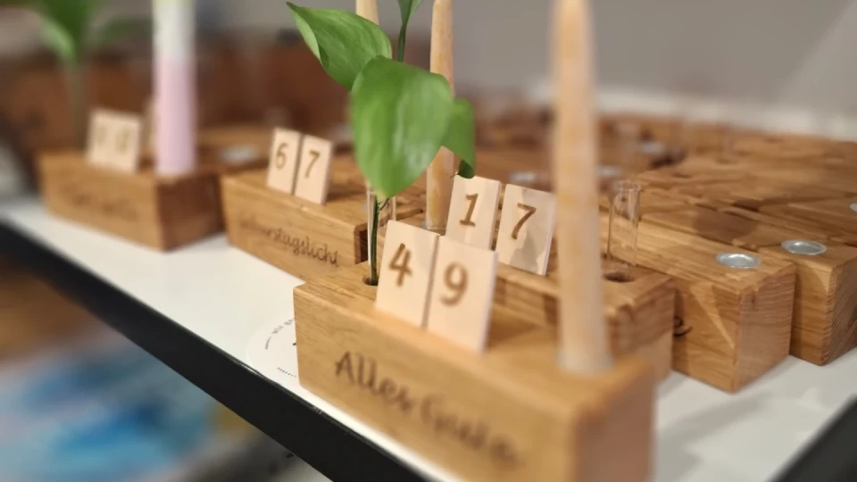 Holzst&auml;nder mit eingefr&auml;stem Text "Allen G&uuml;ten", darauf Holzkl&ouml;tze mit den Zahlen 4, 9, 1, 7 in separaten Schlitzen. Oben sind zwei Holz-Stifte und ein Pflanzenspr&ouml;ssling in einer kleinen Vase integriert. Dahinter &auml;hnliche St&auml;nder mit weiteren Zahlen und Pflanzen.