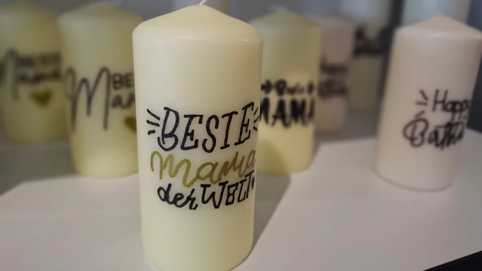 Wei&szlig;e, zylindrische Kerze mit schwarzer und goldener Aufschrift "Beste Mama der Welt", umgeben von weiteren Kerzen mit teils unscharf erkennbaren, &auml;hnlich gestalteten Schriftz&uuml;gen.