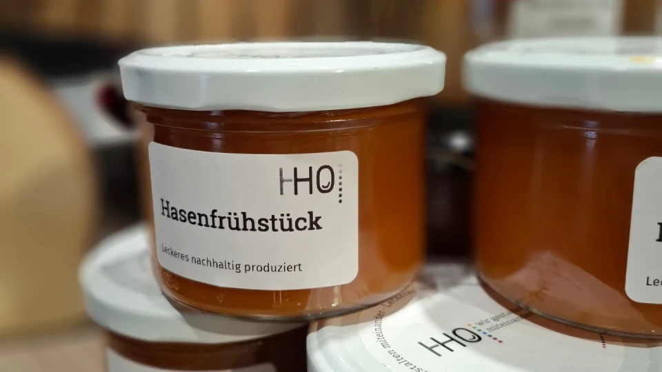 Gl&auml;ser mit wei&szlig;em Deckel und Etikett, auf dem "Hasenfr&uuml;hst&uuml;ck" sowie "Leckeres nachhaltig produziert" steht. Das Glas enth&auml;lt eine orangefarbene Substanz. Im Hintergrund weitere unscharfe Gl&auml;ser und braune Elemente.