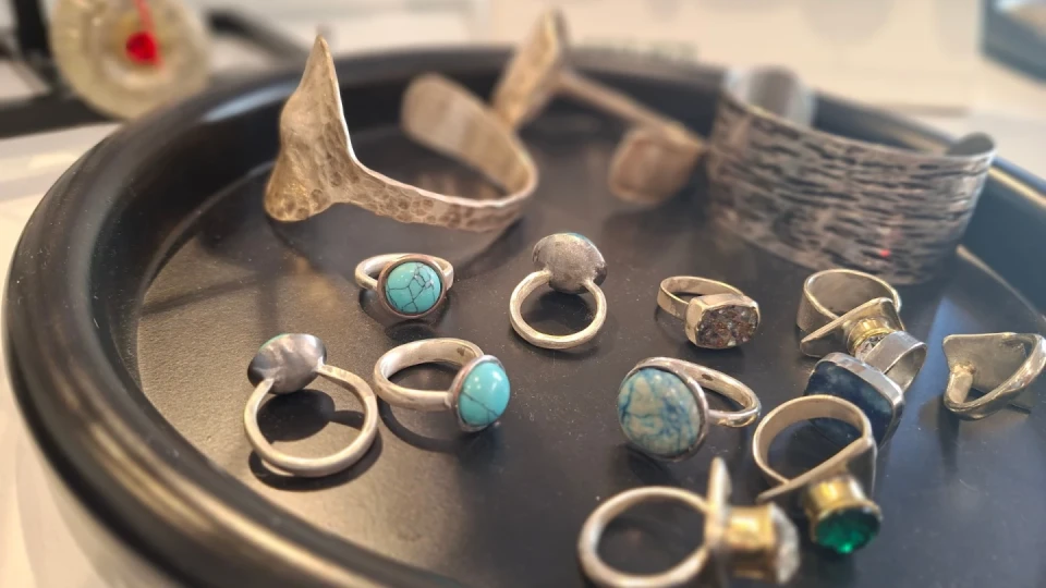 Silberschmuck auf einem schwarzen Tablett: Mehrere Ringe mit Schmucksteinen in Blau, T&uuml;rkis und Transparent sowie schlichte Silberringe. Im Hintergrund zwei breite Armb&auml;nder mit geh&auml;mmertem und strukturiertem Design. Leicht unscharfer Hintergrund.