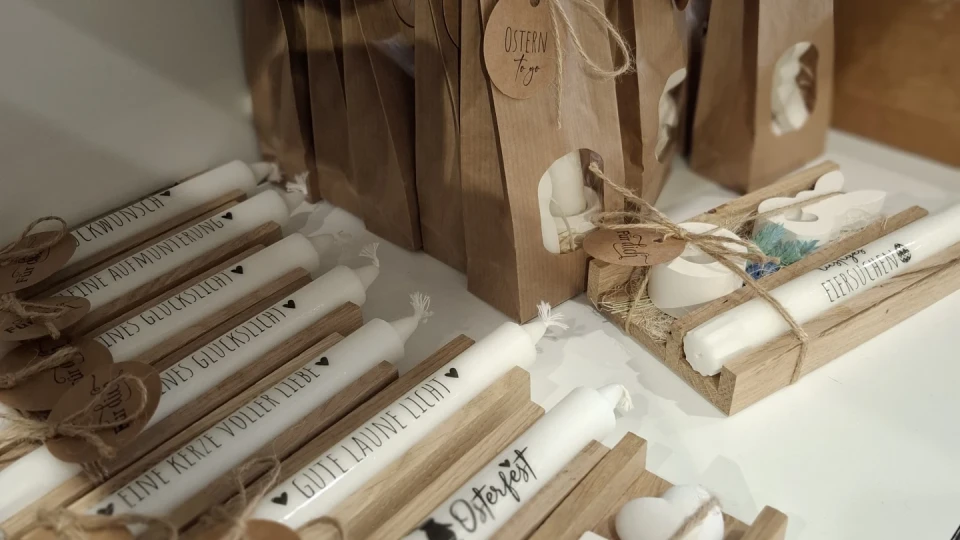 Wei&szlig;e, l&auml;ngliche Kerzen in verschiedenen arrangierten Pr&auml;sentationen. Links Kerzen auf Holzbrettern, teils mit Text wie &bdquo;F&uuml;nf Kekse voller Liebe&ldquo; und &bdquo;Ostern&ldquo; bedruckt, mit runden, braunen Papieranh&auml;ngern aus Bast befestigt. Rechts wei&szlig;e Kerzen in braunen Papiert&uuml;ten, geb&uuml;ndelt und verziert mit Schleifen aus Naturbast.