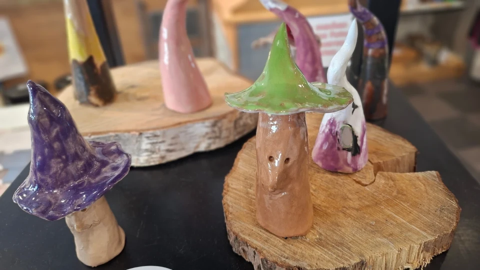 Keramiksammelteller mit dekorativen pilzf&ouml;rmigen Figuren. Eine braun glasierte Figur in der Mitte tr&auml;gt einen gr&uuml;nen Hut mit schr&auml;ger Spitze. Links daneben eine violett glasierte Figur, rechts wei&szlig;e und rosa bemalte Pilzfiguren. Figuren stehen auf runden Holzscheiben. Im Hintergrund verschwommene Einrichtung.