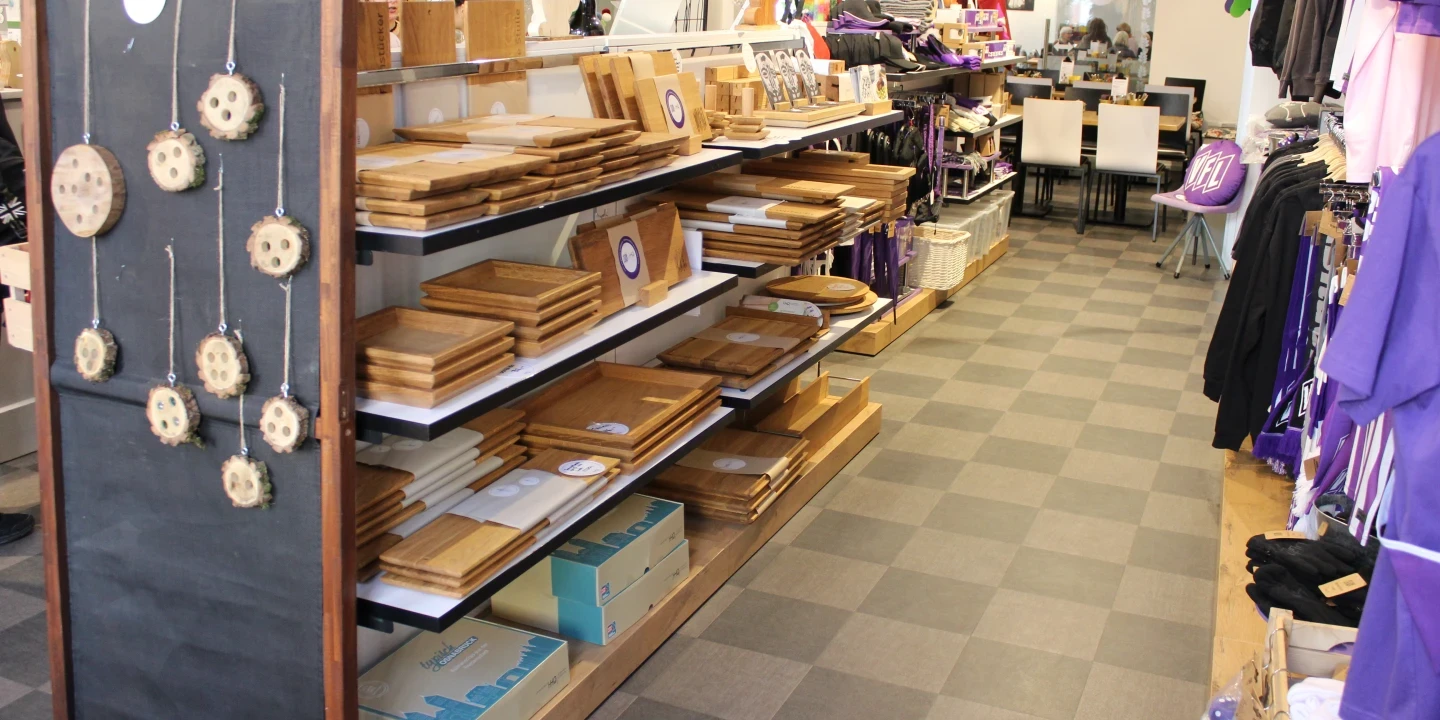 Regale mit gestapelten Holzschneidebrettern und Produktverpackungen, links eine Tafel mit h&auml;ngenden Holzanh&auml;ngern, dahinter Tischgruppe mit wei&szlig;en St&uuml;hlen und dekorativen Artikeln, rechts Kleidung und Accessoires in Lila. Fliesenboden in grau kariertem Muster.