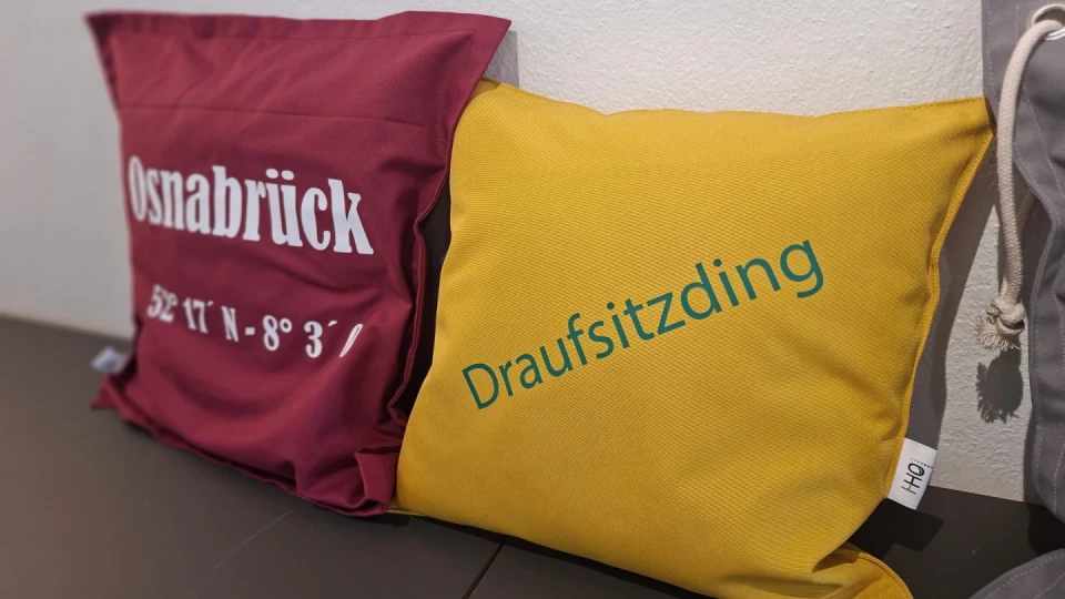 Bordeauxfarbenes Kissen mit wei&szlig;em Schriftzug "Osnabr&uuml;ck 52&deg; 17' N, 8&deg; 3'" links neben einem gelben Kissen mit gr&uuml;nem Aufdruck "Draufsitzding", platziert auf einer dunklen Oberfl&auml;che vor einer wei&szlig;en Wand. Rechts teilweise sichtbar der Kordelzug eines Beutels.