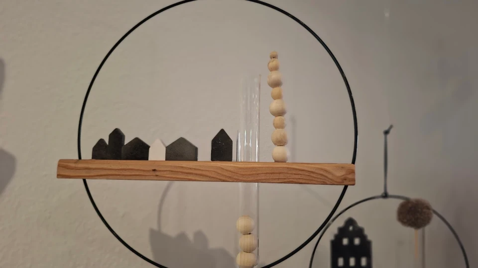 Holzbrett mit kleinen dunkelgrauen, schwarzen und einem wei&szlig;en Miniaturhaus darauf. Rechts steht eine Glasr&ouml;hre mit einer vertikalen Reihe beiger Holzkugeln. Das Arrangement befindet sich in einem schwarzen Metallkreis, der als Rahmen dient. Im unscharfen Hintergrund sind Formen von Dekorationen zu erkennen.