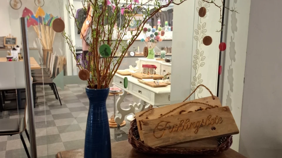 Blau glasierte Vase mit d&uuml;nnen Zweigen darin, dekoriert mit bunten runden Anh&auml;ngern. Davor ein Holzschild mit der Aufschrift "Fr&uuml;hlingsbote" in einer flachen Korbschale. Im Hintergrund ein Raum mit dekoriertem Tisch, St&uuml;hlen und farbenfrohem Dekor auf einem grauen Boden.