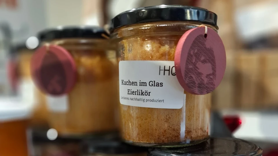 Etikettierter Glasbeh&auml;lter mit hellbraun-goldenem Kuchen und schwarzem Deckel. Auf dem Etikett steht "Kuchen im Glas Eierlik&ouml;r" und "Leckeres nachhaltig produziert". Links daneben unscharf weitere Gl&auml;ser mit &auml;hnlichem Inhalt. Rosa Papieranh&auml;nger am Deckel befestigt.