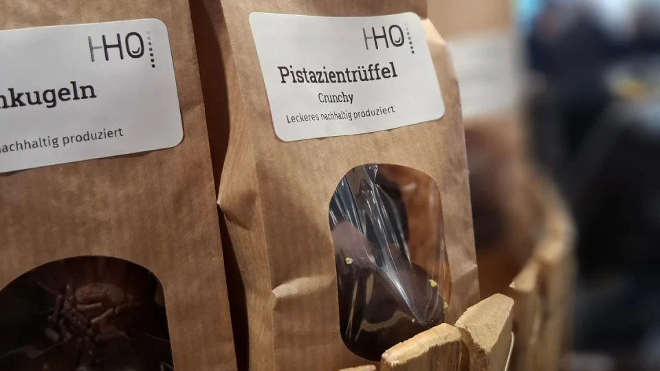 Braune Papiert&uuml;ten mit der Aufschrift "HO! Pistazientr&uuml;ffel Crunchy &ndash; Leckeres nachhaltig produziert", teils durchsichtiges Fenster zeigt dunkle Pralinen. Verpackungen stehen in einem Holzbeh&auml;lter. verschwommener Hintergrund.