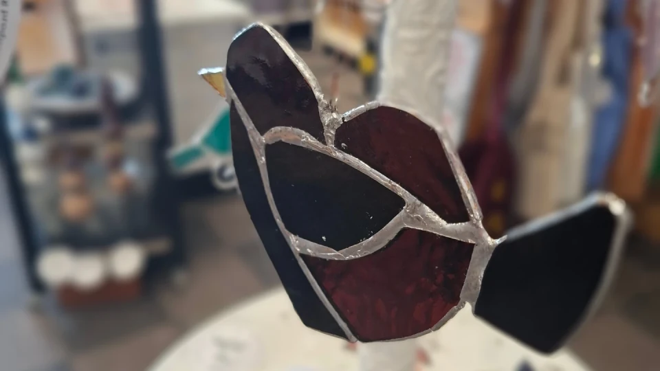 Glasdekor in Form eines stilisierten Vogels mit dunkelroten und schwarzen Glasfl&auml;chen, getrennt durch silberne Metallstreifen. Der Vogel ist links ausgerichtet und h&auml;ngt in einem Innenraum mit unscharfen Regalen und Gegenst&auml;nden im Hintergrund.