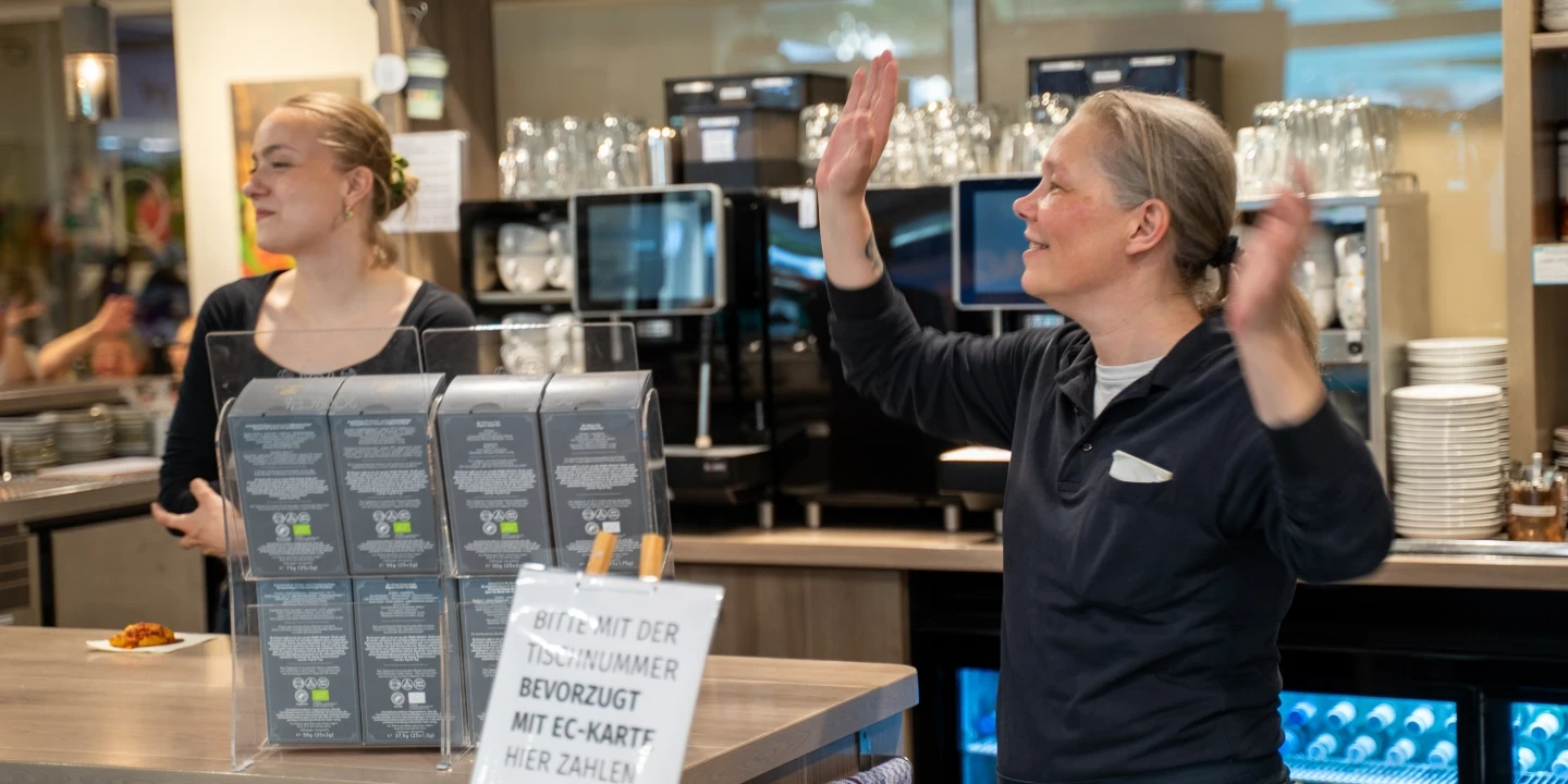 Zwei Personen stehen hinter einer Kaffeetheke in einem Caf&eacute;. Links mehrere Speisekarten auf St&auml;ndern, rechts eine Frau mit erhobenen Armen. Im Hintergrund Kaffeemaschinen, Gl&auml;ser und wei&szlig;e Dekokannen. Im Vordergrund ein Schild mit der Aufschrift &bdquo;Bitte mit der Tischnummer bevorzugt mit EC-Karte hier zahlen&ldquo;.