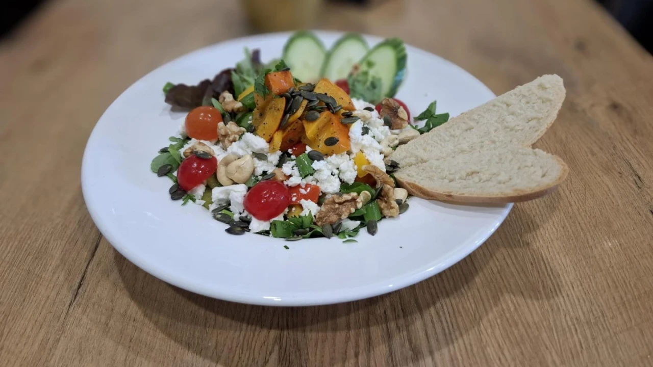 Teller mit Salat aus buntem Gem&uuml;se und Toppings; Rucola, rote Tomaten, K&uuml;rbisw&uuml;rfel, Waln&uuml;sse, Fetak&auml;se und K&uuml;rbiskerne. Beilage aus zwei Wei&szlig;brotscheiben, dahinter Gurkenscheiben und Salatbl&auml;tter. Holzuntergrund.