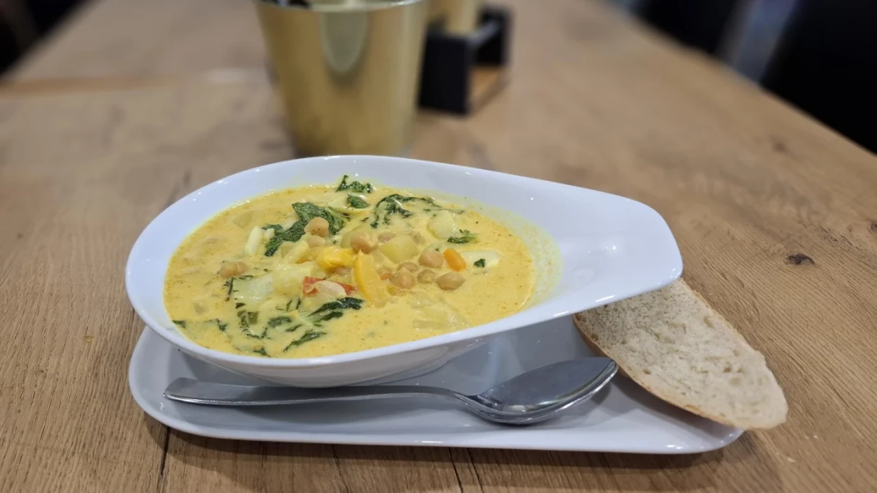 Gelbe Suppe in einem wei&szlig;en ovalen Teller mit Inhaltsstoffen wie Spinatbl&auml;ttern, Gem&uuml;sew&uuml;rfeln und Kichererbsen, serviert auf einem wei&szlig;en rechteckigen Teller mit einem Metalll&ouml;ffel und einer Scheibe hellen Brots. Im Hintergrund ein Tisch aus hellem Holz mit einem goldenen Metallbeh&auml;lter und weiteren Gegenst&auml;nden.
