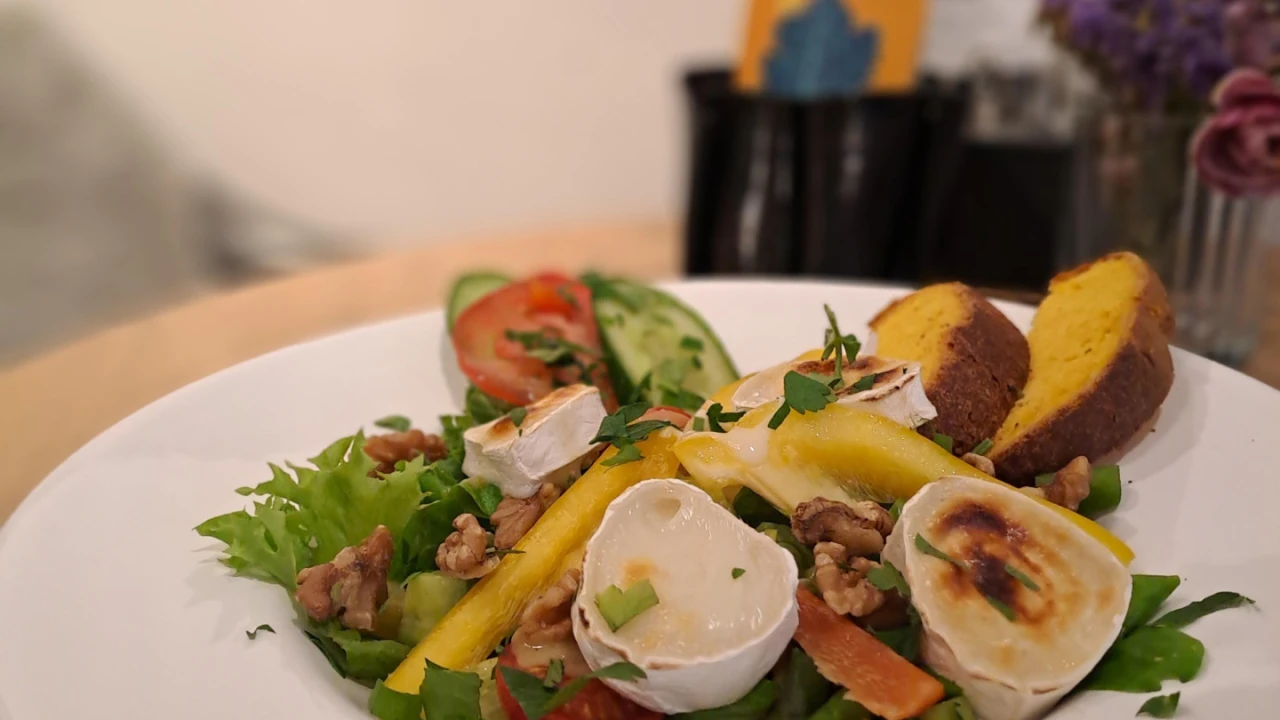 Frische gr&uuml;ne Salatbl&auml;tter mit Cherrytomaten, Gurkenscheiben, gegrilltem Ziegenk&auml;se, gelben Mangostreifen, Waln&uuml;ssen und Kr&auml;utern, serviert auf einem wei&szlig;en Teller. Rechts gebratene Brotscheiben. Im Hintergrund unscharfe Dekoration und violette Blumen.