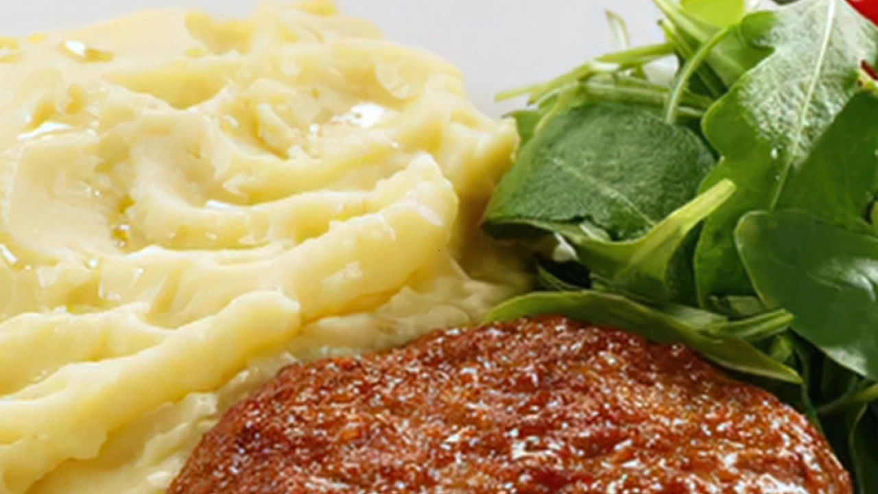 P&uuml;ree aus Kartoffeln mit hellgelber, cremiger Textur links im Bild, daneben ein braun gl&auml;nzendes Hacksteak. Rechts frischer gr&uuml;ner Salat mit hellem Blattwerk. Im Hintergrund rote Tomaten und ein wei&szlig;es Tuch mit hellbraunen Streifen.