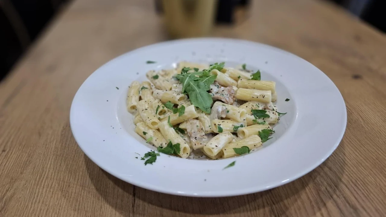 Pasta auf einem wei&szlig;en Teller, serviert mit heller Sahneso&szlig;e, kleinen Fleischst&uuml;cken und frischen Kr&auml;utern, garniert mit Rucola. Der Teller steht auf einem langen, hellbraunen Holztisch, unscharfer Hintergrund mit goldfarbenem Beh&auml;lter und weiteren verschwommenen Objekten.