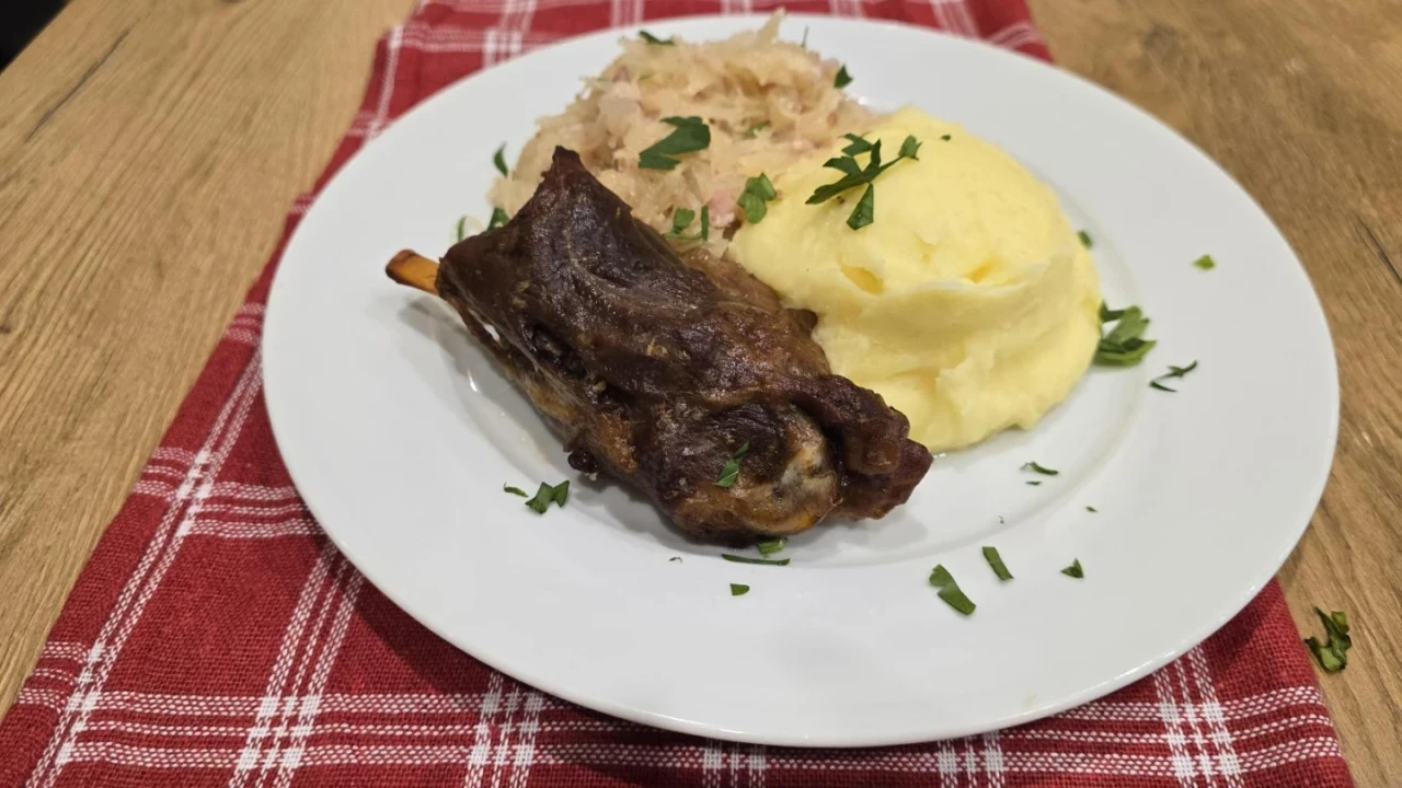 Gegrilltes Fleischst&uuml;ck mit Knochen, daneben gelber Kartoffelbrei und beige-graues Sauerkraut, serviert auf einem wei&szlig;en Teller. Mit gr&uuml;ner Petersilie garniert, auf rotem, kariertem Stofftischl&auml;ufer und holzfarbener Tischoberfl&auml;che arrangiert.