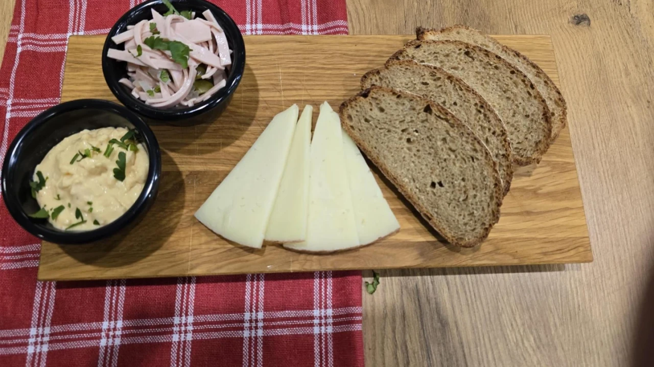 Holzbrett mit vier Scheiben braunem Vollkornbrot, f&uuml;nf dreieckigen K&auml;sescheiben in der Mitte, einer schwarzen Schale mit Aufstrich garniert mit Petersilie und einer weiteren schwarzen Schale mit geschnittenem Aufschnitt, dekoriert mit Petersilie. Unterlage aus rot-wei&szlig; kariertem Stoff auf einem Holztisch.