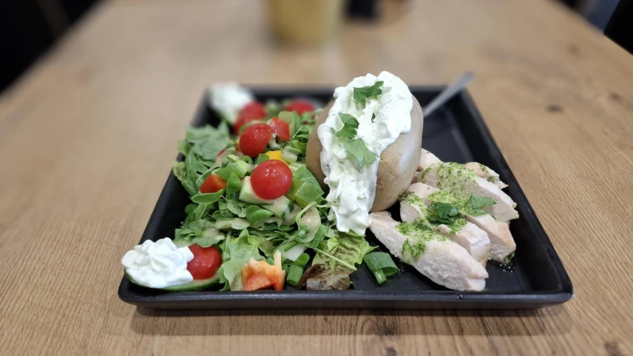 Schwarzer rechteckiger Teller mit gegrillten H&auml;hnchenst&uuml;cken, garniert mit gr&uuml;nen Kr&auml;utern, einer gebackenen Kartoffel mit wei&szlig;em Kr&auml;uterdip, einer Portion gemischten Salats aus Blattsalat, Kirschtomaten und Paprikast&uuml;cken sowie kleinen Klecksen mit Kr&auml;uterdip. Teller steht auf einem braunen Holztisch.