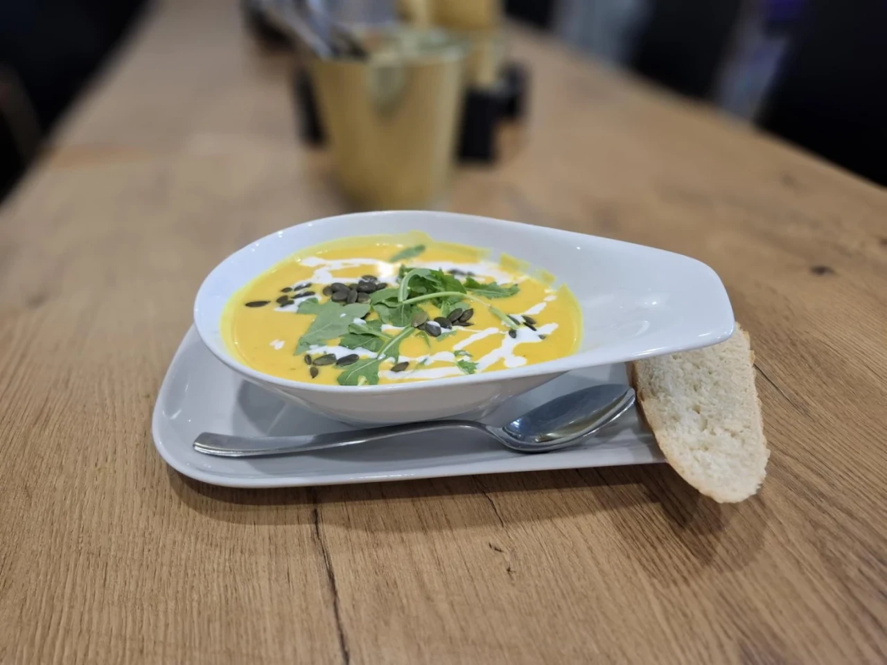 Wei&szlig;e, ovale Schale mit orangefarbener Cremesuppe, dekoriert mit K&uuml;rbiskernen, gr&uuml;nen Kr&auml;utern und einem Sahne-Muster, auf einem wei&szlig;en rechteckigen Teller mit einem silbernen L&ouml;ffel und einer Scheibe Wei&szlig;brot. Holztisch im Hintergrund.