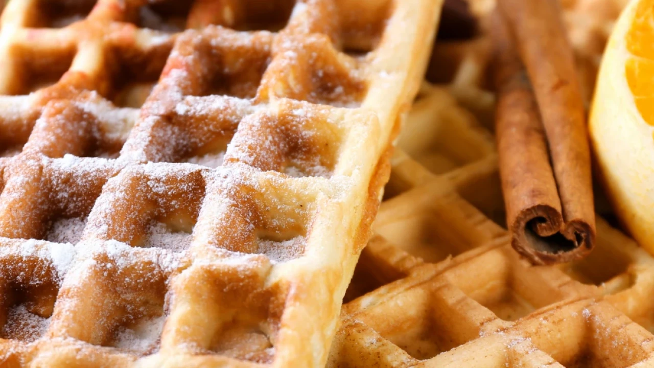 Zwei goldbraune Waffeln auf einem h&ouml;lzernen Untergrund, best&auml;ubt mit Puderzucker, dekoriert mit zwei Zimtstangen, einem frischen Minzblatt und einem St&uuml;ck Orangenscheibe am rechten Rand.