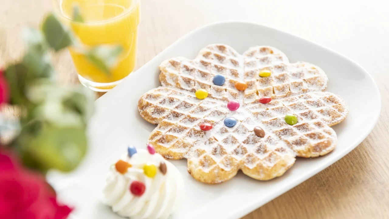 Herzf&ouml;rmige Waffel auf einem wei&szlig;en rechteckigen Teller, bestreut mit Puderzucker und dekoriert mit bunten Schokolinsen, daneben ein kleiner Klecks Schlagsahne mit weiteren Schokolinsen garniert. Im Hintergrund ein Glas mit orangem Saft und unscharfe rote und gr&uuml;ne Blumendetails.