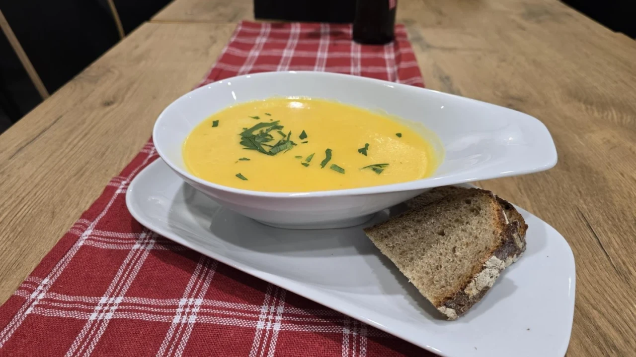 Wei&szlig;e ovale Schale mit gelber Suppe, garniert mit gr&uuml;nen Kr&auml;utern, auf l&auml;nglichem wei&szlig;en Teller. Rechtsseitig ein St&uuml;ck braunes Brot mit Kruste. Roter Tischl&auml;ufer mit wei&szlig;em Karomuster auf hellem Holztisch, Details im Hintergrund unscharf.