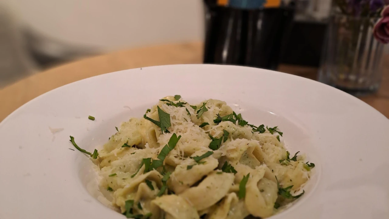 Teller mit gr&uuml;ner Pasta in heller Sauce, garniert mit gehackter Petersilie und geriebenem K&auml;se. Im Hintergrund ein Tisch mit einer Vase mit lilafarbenen Blumen und aufgestellten Karten.