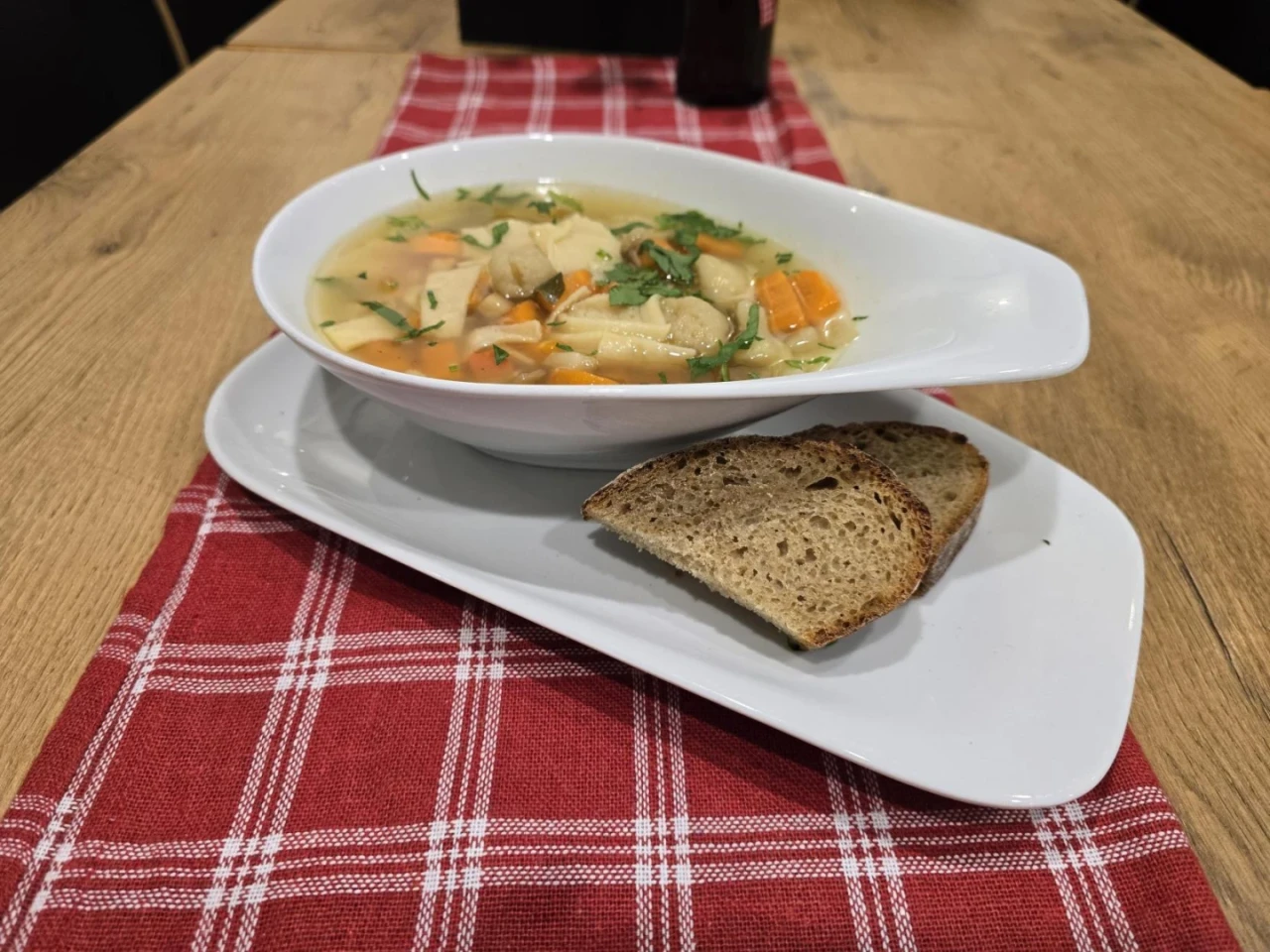 Wei&szlig;e, ovale Suppenschale mit Gem&uuml;se- und Nudelsuppe, garniert mit Kr&auml;utern, auf wei&szlig;em Teller; daneben zwei Scheiben dunkles Brot. Teller auf rot-wei&szlig; kariertem Stoffl&auml;ufer auf hellem Holztisch.