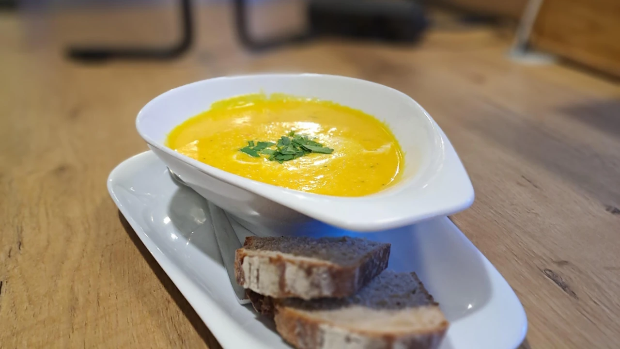 Wei&szlig;e, ovale Schale mit gelber, cremiger Suppe, garniert mit gr&uuml;nen Kr&auml;utern, auf einer l&auml;nglichen wei&szlig;en Untertasse platziert. Daneben liegen zwei Scheiben dunkles Brot mit heller Kruste. Der Untergrund besteht aus einer Holzoberfl&auml;che, im Hintergrund unscharfe Details eines Raums.