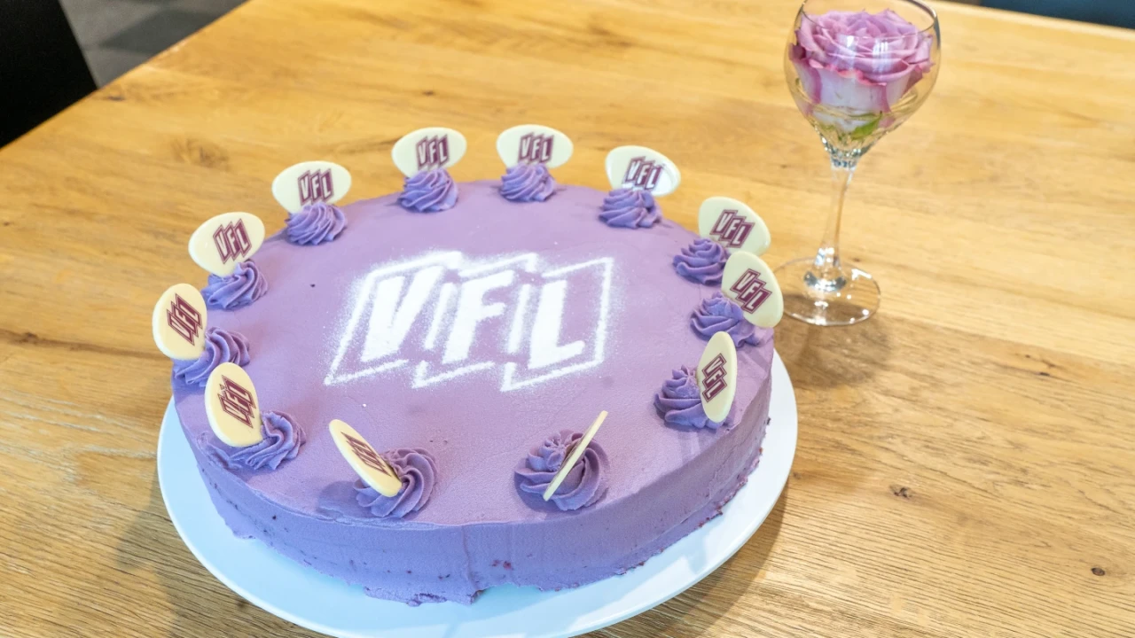 Lila Torte auf wei&szlig;em Teller auf einem Holztisch, dekoriert mit violetten Creme-Tupfen und wei&szlig;en Schokoladenscheiben mit dem lila "VfL"-Logo; mittig ebenfalls das "VfL"-Logo aus Zuckerguss. Rechts ein Weinglas mit einer eingearbeiteten lila-wei&szlig;en Rose.