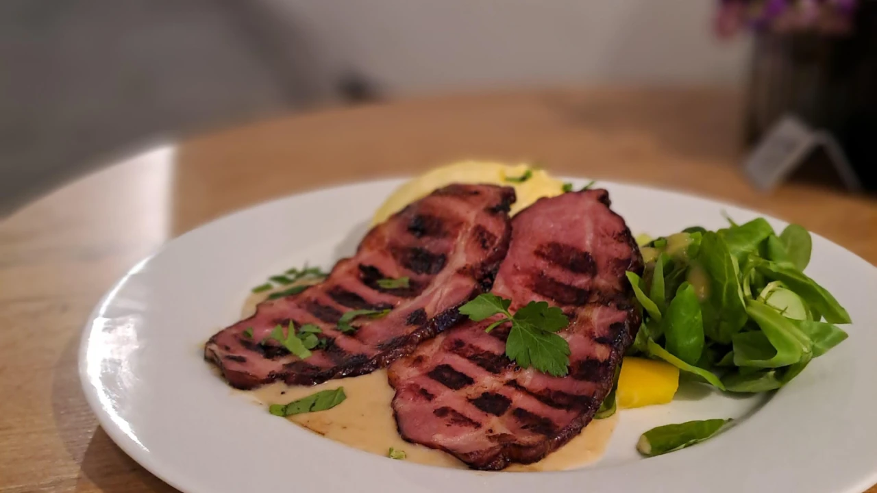 Wei&szlig;er Teller mit drei gegrillten, braun-rosa Fleischscheiben mit Grillstreifen, angerichtet auf cremiger heller Sauce, daneben ein gelber Gem&uuml;sew&uuml;rfel und ein kleiner Haufen gr&uuml;ner Salatbl&auml;tter. Im Hintergrund ein verschwommener Tisch mit Blumen.