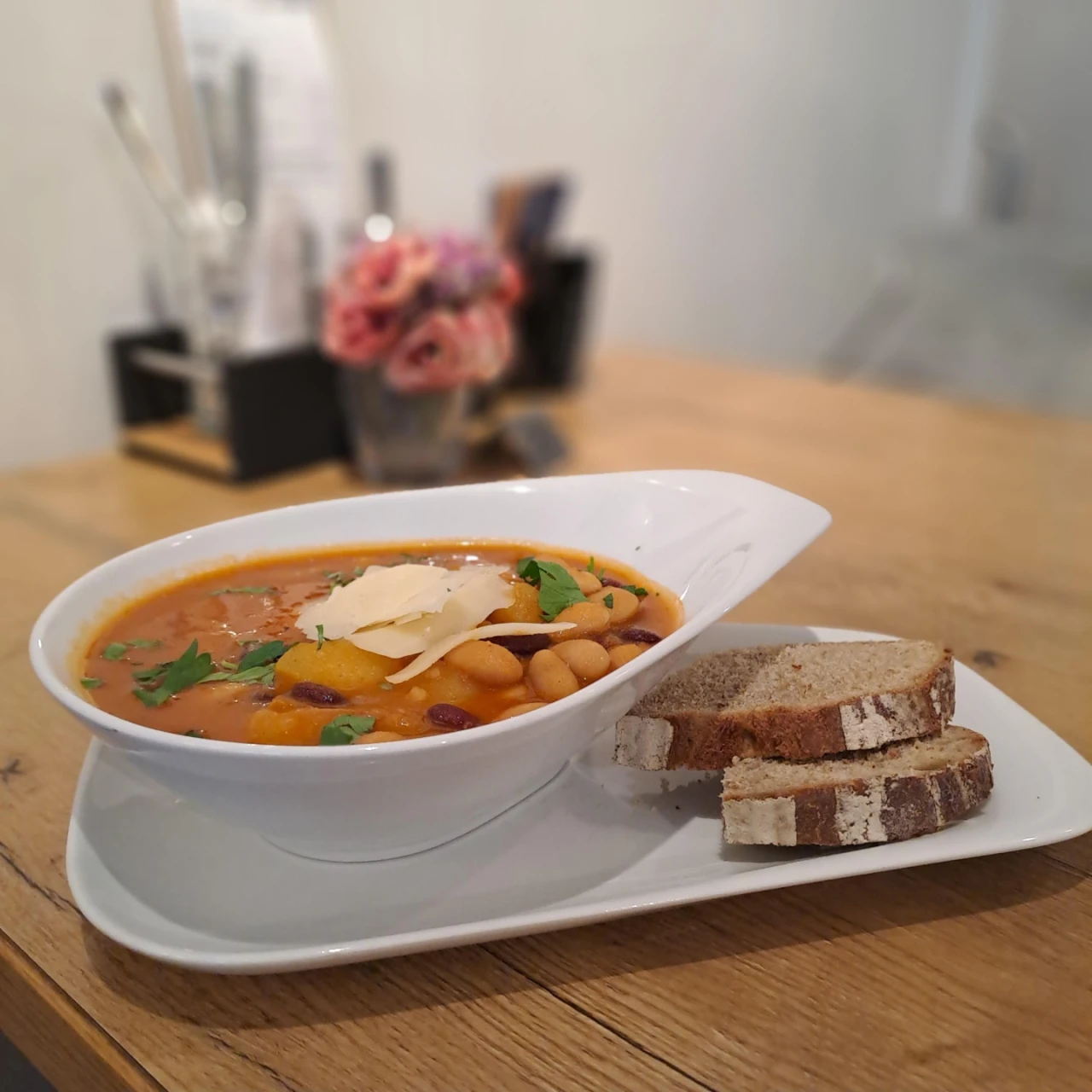 Wei&szlig;e, ovale Sch&uuml;ssel mit einer dickfl&uuml;ssigen Gem&uuml;sesuppe, garniert mit gehobeltem K&auml;se, Petersilie, Kichererbsen und roten Kidneybohnen, serviert auf einem wei&szlig;en Teller mit zwei Scheiben braunem Brot. Im Hintergrund unscharfe Tischdekoration mit pinken Bl&uuml;ten und Besteckhalter auf einem Holztisch.