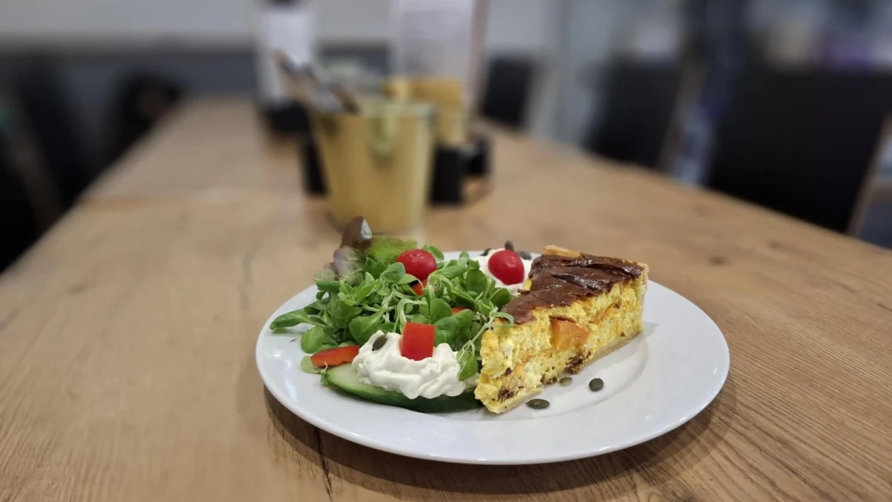 Wei&szlig;e Porzellanteller auf Holztisch mit herzhaftem St&uuml;ck Quiche, daneben frischer Salat aus Rucola und anderen gr&uuml;nen Bl&auml;ttern, garniert mit roten Tomatenst&uuml;cken und Sahneh&auml;ubchen, dekoriert mit K&uuml;rbiskernen. Verschwommene Hintergrundelemente einer Caf&eacute;-Inneneinrichtung und ein Besteckhalter mit Utensilien.