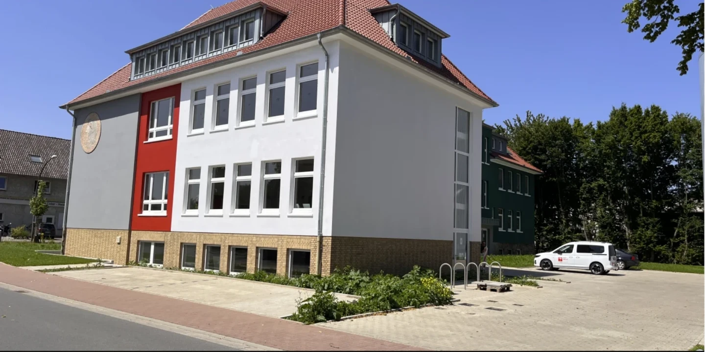 Eckgeb&auml;ude mit wei&szlig;er Fassade und rotem Fassadenabschnitt, gelber Sockelbereich. Vier Fensterreihen mit gro&szlig;en Glasfenstern. Rotes Ziegeldach mit mehreren Gauben. Parkplatz rechts mit zwei Autos, gr&uuml;ne B&auml;ume im Hintergrund, Fahrradst&auml;nder, Fu&szlig;weg und Stra&szlig;e im Vordergrund. Blauer Himmel.