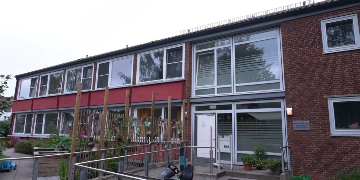 Zweigeschossiges Geb&auml;ude mit roter und roter Backsteinfassade. Links Holzlatten mit Pflanzen dazwischen. Rechts gro&szlig;e Glasfenster und Eingang mit Rampe daneben. Flache D&auml;cher und niedriges Gel&auml;nder.