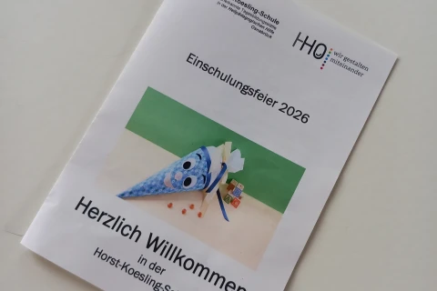 Faltblatt auf wei&szlig;em Tisch mit der Aufschrift: "Horst-Koesling-Schule", "Einschulungsfeier 2026", und "Herzlich Willkommen in der Horst-Koesling-Schule". Darauf ein Bild einer blau-wei&szlig; gemusterten Schult&uuml;te mit Gesicht, gef&uuml;llt mit kleinen Geschenken, daneben Buchstabenw&uuml;rfel und orangefarbene S&uuml;&szlig;igkeiten. Logo oben rechts: "HHO &ndash; wir gestalten miteinander".