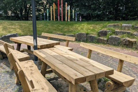 Holztische und -b&auml;nke mit rustikalem Design unter einem zusammengefalteten gelben Sonnenschirm auf einer gepflasterten Fl&auml;che. Im Hintergrund eine gr&uuml;ne Rasenfl&auml;che mit gro&szlig;en Steinen und bemalten Holzpf&auml;hlen in bunten Farben, umgeben von hohen B&uuml;schen und B&auml;umen.