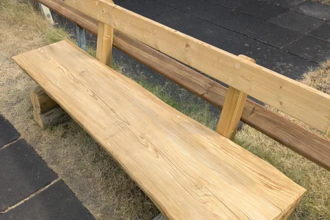 Holzbank aus hellem, naturbelassenem Holz auf einem Spielplatz mit schwarzen Gummimatten als Bodenbelag. Im Hintergrund ein gr&uuml;nes, metallisches Spielger&auml;t mit schwarzen Fl&auml;chen sowie Holzb&auml;nke und ein blauer Plastikstuhl auf Rasen.