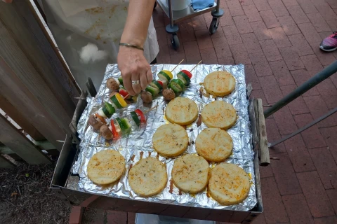 Person h&auml;lt zwei bunte Grillspie&szlig;e aus Gem&uuml;se und Fleisch &uuml;ber einer mit Aluminiumfolie bedeckten Grillfl&auml;che. Darauf liegen neun runde Brotscheiben, goldbraun gebacken. Der Boden besteht aus roten Pflastersteinen, rechts ein Schuh und ein Metallgestell sichtbar.