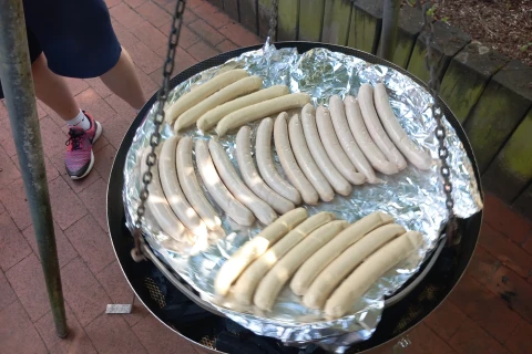 Mehrere rohe, helle Bratw&uuml;rste auf runder Grillfl&auml;che mit Aluminiumfolie bedeckt, aufgeh&auml;ngt an Metallketten. Im Hintergrund Backsteindöner-Boden, ein Bein mit kurzem blauen Hosenstoff, wei&szlig;en Socken und pink-schwarzen Turnschuhen erkennbar. Holzbalken und gr&uuml;ne Beetumrandung sichtbar.