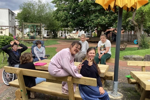 Kinder und Jugendliche sitzen und stehen an Holzsitzb&auml;nken in einem Hof mit gepflastertem Boden, Rasen und B&auml;umen. Eine gelbe Schirmspitze ragt ins Bild. Im Hintergrund sind Spielger&auml;te, Z&auml;une, Geb&auml;ude und ein Baum erkennbar. Einige Personen sitzen l&auml;chelnd, zwei Kinder machen Handgesten.