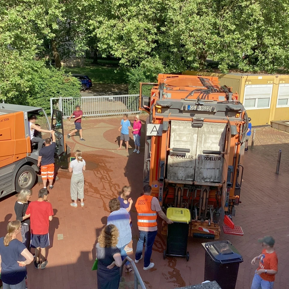 Personen in Freizeitkleidung und orangefarbenen Arbeitswesten stehen auf einem gepflasterten Hof nahe zweier orangefarbener M&uuml;llfahrzeuge. Eine Person schiebt einen gelben M&uuml;llcontainer zum Fahrzeug. Im Hintergrund gr&uuml;ne B&auml;ume, ein Zaun, Containergeb&auml;ude und geparkte Autos.