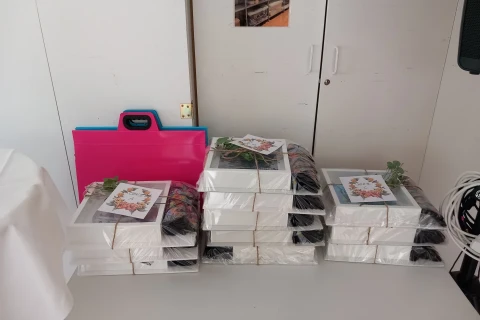 Gestapelte wei&szlig;e Boxen in Plastikverpackung, sortiert in drei Gruppen auf einer Tischplatte. Jede Box hat eine Karte mit floralen Motiven darauf. Links ein pink-blauer Ordner, rechts wei&szlig;e Kabel und technische Ger&auml;te. Dahinter ein wei&szlig;er Schrank mit geschlossenenen T&uuml;ren.