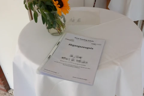 Runder Stehtisch mit wei&szlig;em Tischtuch, darauf eine Glasvase mit Sonnenblume und gr&uuml;nen Zweigen, daneben ein Abgangszeugnis der "Horst-Koesling-Schule", Schrift teilweise unleserlich. Hintergrund mit wei&szlig;en W&auml;nden und farbigen Gegenst&auml;nden, darunter Taschen. Boden mit gefliesten Steinplatten.