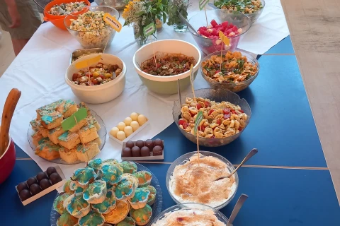 Buffettisch auf einer langen, blauen Oberfl&auml;che mit diversen Speisen: vorn zwei Sch&uuml;sseln mit Dessert und blauen Muffins mit Verzierung. Links Kuchenst&uuml;cke, rechts Salatsch&uuml;sseln mit Gem&uuml;se, Pasta und Obst. In der Mitte Blumenarrangement. Im Hintergrund Brotkorb und Flaschen. Personen stehen seitlich.