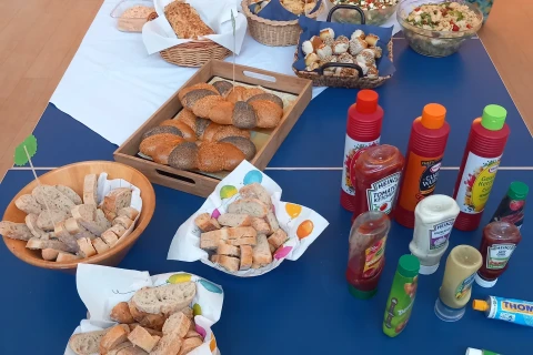 Tisch mit verschiedenen Speisen und Getr&auml;nken auf einer blauen Tischdecke. Links Schalen mit Brotst&uuml;cken, Br&ouml;tchen in K&ouml;rben, in der Mitte Salate und dekorative Blumen. Rechts verschiedene So&szlig;enflaschen, darunter Ketchup, Mayonnaise und Kr&auml;uterso&szlig;en. Menschen im Hintergrund.