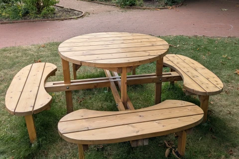 Runder Holztisch mit vier geschwungenen, halbrunden Holzb&auml;nken, die fest mit dem Tisch verbunden sind, auf gr&uuml;nem Rasen. Im Hintergrund ein gepflasterter Weg, gr&uuml;ne B&uuml;sche, ein blau gestrichener Holzverschlag und ein bemalter Zaun vor einem &auml;lteren Geb&auml;ude.