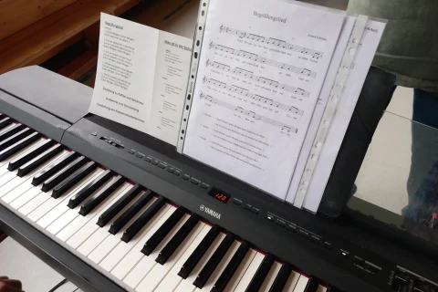 Schwarzes Yamaha-Digitalpiano mit wei&szlig;-schwarzen Tasten, darauf aufgeklappte Notenbl&auml;tter in Klarsichth&uuml;llen, darunter ein &bdquo;Begr&uuml;&szlig;ungslied&ldquo; mit Noten und Text auf hellem Papier. Links neben den Noten weitere Bl&auml;tter mit Texten. Im Hintergrund eine Holzbank und eine Person seitlich im Bild.